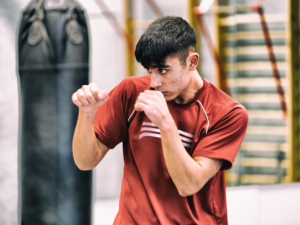 Junger Mann in rotem Trainingsshirt übt Boxbewegungen vor einem Boxsack in einem Trainingsraum.